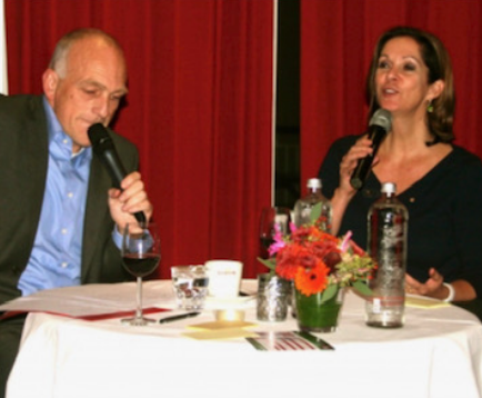 Peter de Bruin interviewt een gast met microfoon, warm avondlicht en rood gordijn